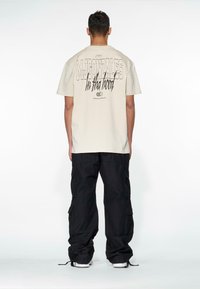 Beige cotton t-shirt with bold black text on the back, worn with loose-fitting black cargo pants. Simple design, relaxed fit, and minimal detailing.