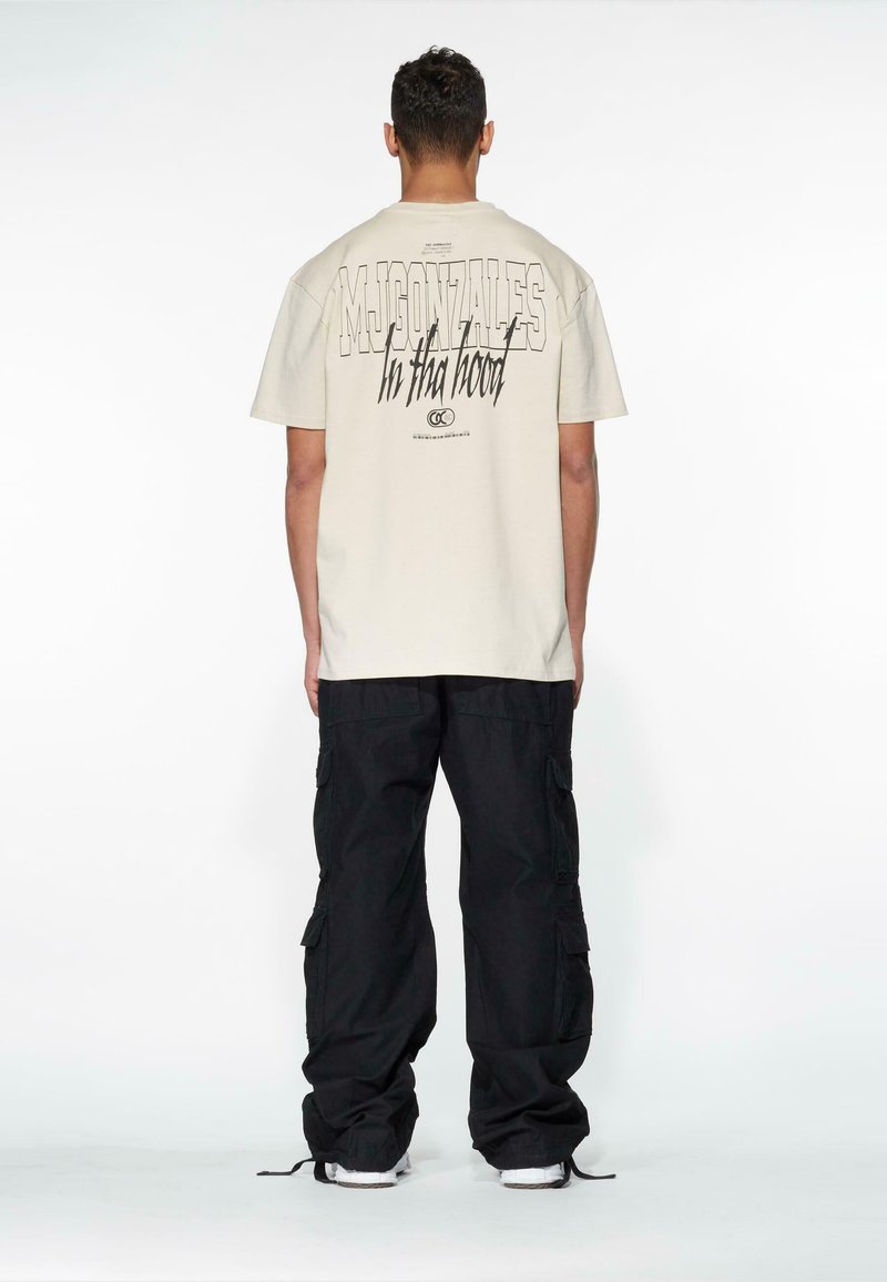 Beige cotton t-shirt with bold black text on the back, worn with loose-fitting black cargo pants. Simple design, relaxed fit, and minimal detailing.