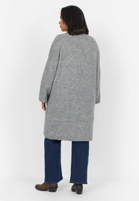 Manteau oversized en tricot gris avec une silhouette droite, des manches longues et une coupe mi-longue, associé à un jean évasé bleu et des bottines brunes.