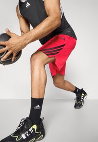 Mand i sort ærmeløs skjorte og røde shorts dribler med en sort basketball, iført sorte Adidas-sokker og sorte træningssko.