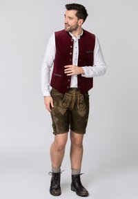 Burgunderfarbener Samt-Westen über einem weißen Hemd, kombiniert mit braunen knie-langen Shorts mit aufwändiger Stickerei und schwarzen Lederstiefeln.