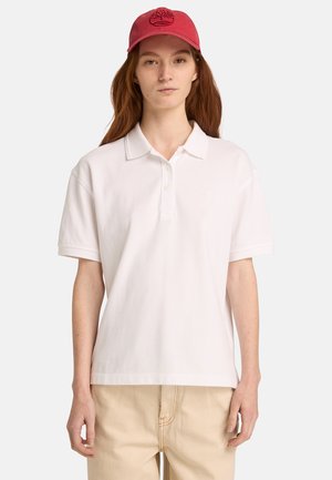 Polo en coton blanc avec une patte de boutonnage à trois boutons et des manches courtes, associé à un pantalon beige clair. Une casquette rouge arbore un logo brodé.