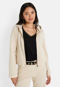 Sudadera beige con cremallera y cordones, de textura suave y con bolsillos frontales, llevada sobre una camiseta negra de cuello en V y pantalones beige claros, complementada con un cinturón.