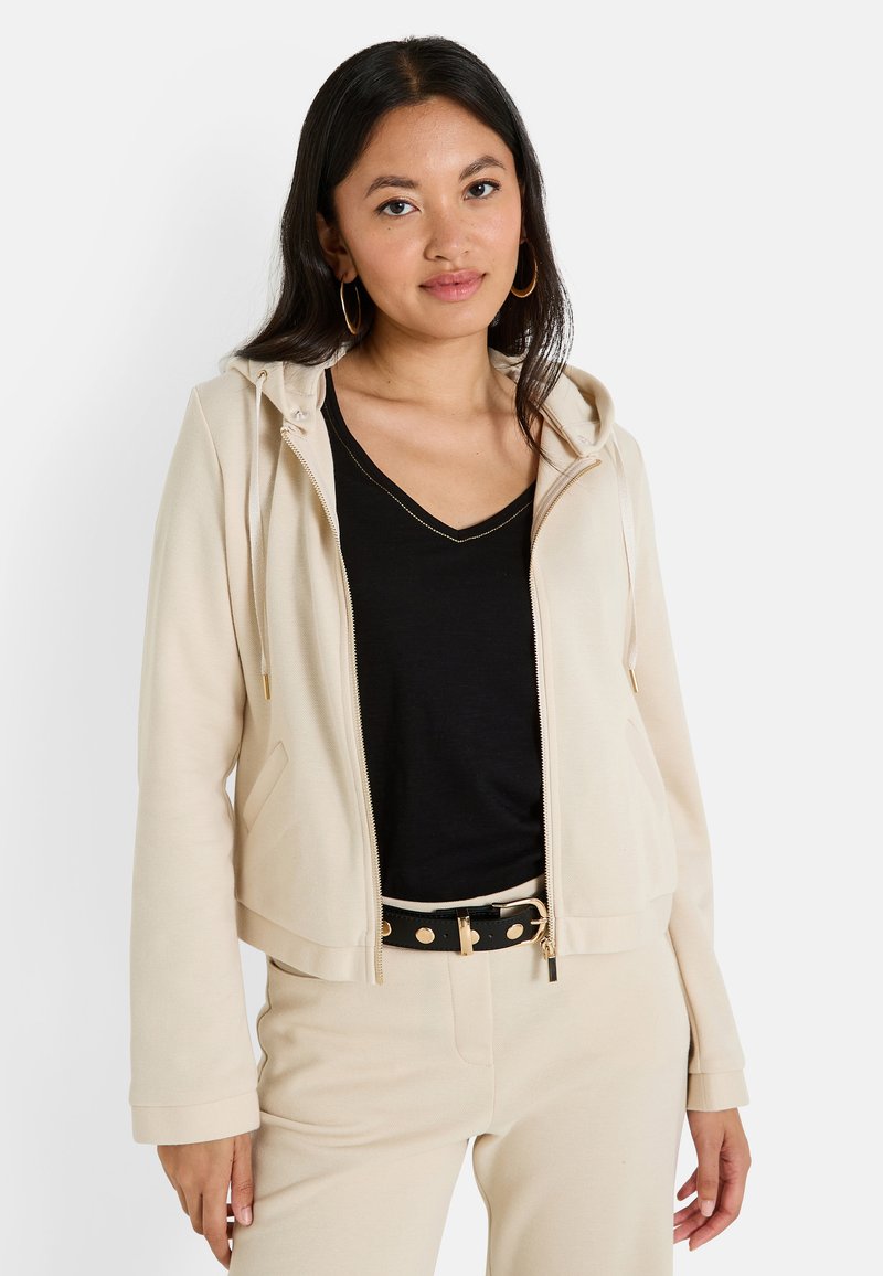 Sudadera beige con cremallera y cordones, de textura suave y con bolsillos frontales, llevada sobre una camiseta negra de cuello en V y pantalones beige claros, complementada con un cinturón.