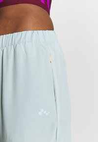 Light blue athletic shorts with an elastic waistband, featuring a small logo in white and an orange accent on the pocket. Smooth texture.