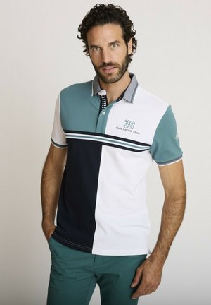 Homme aux cheveux bouclés portant un polo bicolore en bleu sarcelle, marine et blanc, associé à un pantalon bleu sarcelle, debout sur un fond uni.