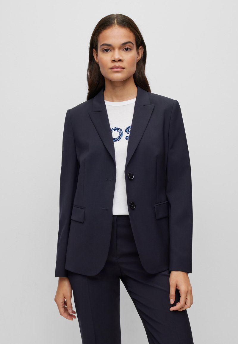 Blazer navy in un design sartoriale, con rever a punta, due tasche frontali e due bottoni, indossato sopra una camicia bianca con scritte blu.