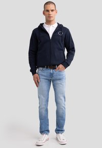 Mees seisab seljas tumesinine lukuga hupsooder, valge polo särk, hele sinised teksad ja valged tossud üle ühetaolise tausta.