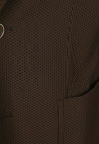 Brauner strukturierten Stoff mit einem Wabenmuster, der eine sichtbare Tasche und Nahtdetails aufweist. Die Knöpfe sind metallisch und sorgen für einen Kontrast.