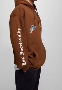 Sudadera marrón con un bolsillo frontal. Presenta un diseño bordado en azul y blanco de un ángel y "Los Angeles City" en blanco en la manga.