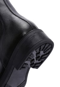 Bottine en cuir noir à semelle en caoutchouc. Présente un motif de semelle texturé et un dessus lisse avec des coutures visibles le long des joints.