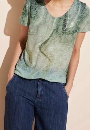 Persoon die een groen shirt met korte mouwen en patroon draagt met tekst en ontwerp op de borst, gecombineerd met donkerblauwe spijkerbroek en handen in de zakken.