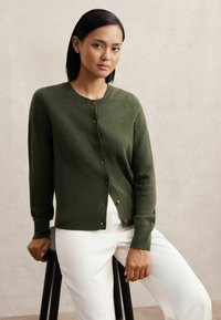 Olíva zöld kardigán, puha kötött anyagból, kerek nyakkivágással, gombos elülső résszel és bordás mandzsettákkal. Fehér nadrág felett viselve.