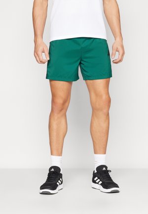 Man draagt groene sportshorts, witte crew-sokken en zwarte Adidas sneakers, staand tegen een effen achtergrond.
