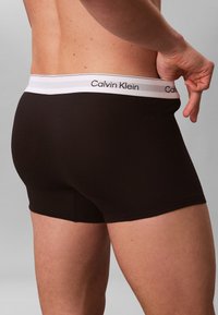 Męski tors i górna część ud w czarnych bokserkach Calvin Klein z białym pasem, ręka lekko ciągnie pas na zewnątrz.
