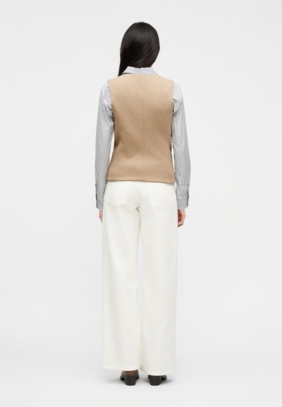 Γυναίκα με μακριά σκούρα μαλλιά, που φοράει μπεζ γιλέκο πάνω από ριγέ πουκάμισο και φαρδιά λευκά παντελόνια, στέκεται γυρισμένη με την πλάτη προς το απλό φόντο.