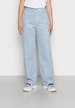 Person trägt hellblaue, weit geschnittene Cordhose, weiße Sneakers mit schwarzen Details und ein weißes kurzärmeliges Oberteil, das in die Hose gesteckt ist.