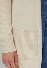 Mano poggiata vicino al polsino rigato e alla tasca rettangolare di un maglione beige lavorato a maglia, con il denim blu visibile sullo sfondo.