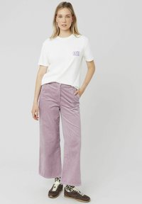 T-shirt en coton blanc avec un graphisme floral violet, associé à un pantalon en velours côtelé large de couleur violet clair. Baskets marron avec des lacets contrastants.