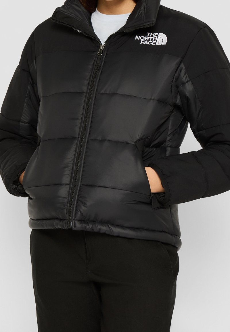 Personne portant une veste matelassée noire The North Face avec une fermeture éclair à l'avant et les mains dans les poches, sur un fond blanc.