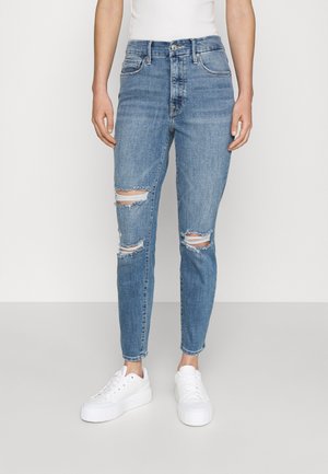 Personne portant un jean skinny déchiré bleu clair avec des trous effilochés aux genoux, des baskets blanches à plateforme et un haut blanc sans manches.