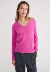 V-ausschnitt, langärmeliger Pullover in leuchtendem Pink aus weichem Material; gerippte Details an den Bündchen und am Saum; getragen mit dunkelgrauen Hosen.