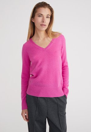 V-ausschnitt, langärmeliger Pullover in leuchtendem Pink aus weichem Material; gerippte Details an den Bündchen und am Saum; getragen mit dunkelgrauen Hosen.