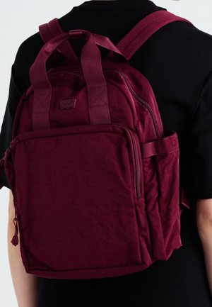 Person trägt einen dunkel weinroten Rucksack mit mehreren Fächern und gepolsterten Schultergurten über einem schwarzen Hemd.