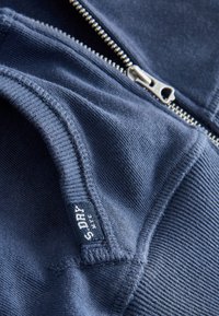 Gros plan d'un sweat à capuche bleu foncé zippé montrant des coutures détaillées et une petite étiquette en tissu avec le texte de la marque.