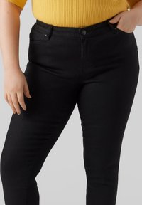 Jeans negros ajustados con una cinturilla estándar, bolsillos delanteros y tela texturizada. Combinados con una blusa amarilla de canalé, mostrando un atuendo informal.