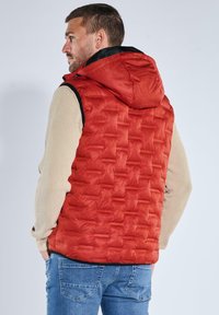 Rote gesteppte Steppweste mit Kapuze und schwarzen Akzenten, getragen über einem beigen Pullover, darunter sind blaue Jeans sichtbar.