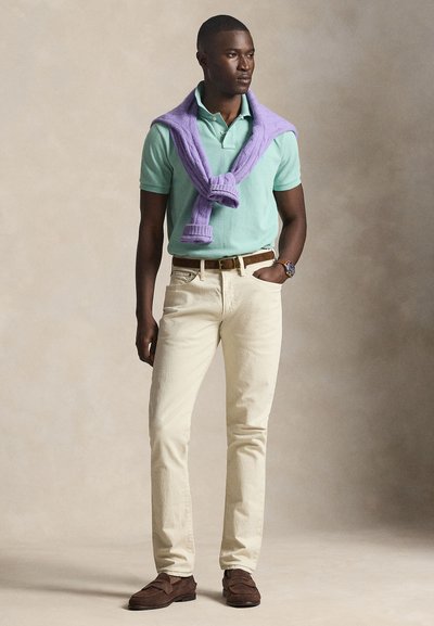 Άνδρας που φοράει ανοιχτοπράσινο πόλο, μπεζ παντελόνι, καφέ ζώνη και παπούτσια, με μωβ πουλόβερ περασμένο στους ώμους, στέκεται με το ένα χέρι στην τσέπη.