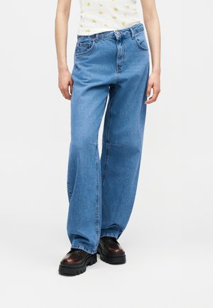 Személy világoskék, széles szárú farmert, fehér, sárga virágmintás inget és fekete, vastag talpú cipőt visel, fehér háttér előtt.