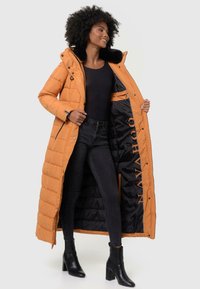 Manteau d'hiver long et orange en duvet avec un col en fourrure synthétique, doté d'une fermeture éclair et d'une fermeture à bouton. Intérieur noir avec logo. Porté sur un jean foncé et des bottines.