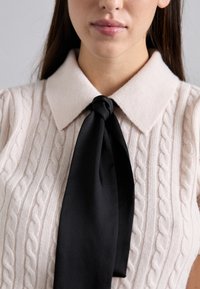 Pull en tricot de couleur beige clair avec des motifs en câble, présentant un col structuré et un nœud en satin noir au niveau du cou.