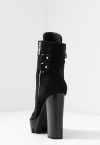 Černá semišová kotníková bota s vysokým masivním podpatkem, vybavená bočním zipem a dvojitými přezkami pro ozdobu. Hladký povrch a elegantní design.