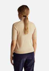 Donna con coda di cavallo castano chiaro che indossa un maglione a collo alto corto beige e pantaloni in velluto blu scuro, di spalle.