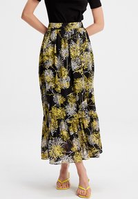 Jupe longue noire avec un imprimé floral jaune et blanc, design plissé, tissu fluide et ourlet superposé. Associée à des sandales jaunes.