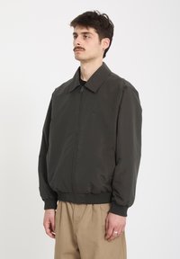 Homme moustachu portant une veste zippée vert foncé et un pantalon beige, debout devant un fond blanc uni.