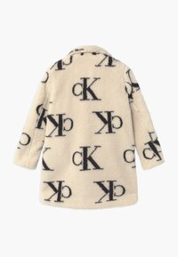 Faux fur coat in cream, featuring a black logo pattern. Long sleeves, collared design, and a relaxed fit with soft texture.