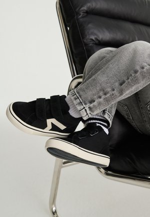 Child wearing black sneakers with white soles and velcro straps, gray jeans, and striped socks sitting on a black leather chair.