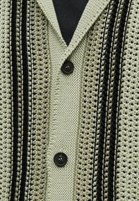 Κοντινή λήψη πλεκτής ζακέτας με υφασματικά σχέδια, κάθετες μαύρες και μπεζ ρίγες και δύο μαύρα κουμπιά κουμπωμένα πάνω από ένα μαύρο πουκάμισο.