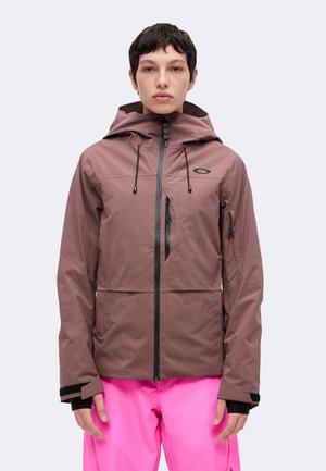 Personne aux cheveux courts et foncés porte une veste à capuche marron avec une fermeture éclair noire et un pantalon rose vif, debout devant un arrière-plan clair uni.