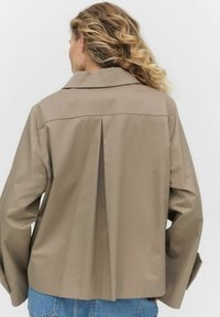 Beige, rakenteellinen takki, jossa on kärkipaita, laskosyksitys takana ja pitkät hihat. Materiaalin pinta näyttää sileältä ja hieman kiiltävältä.