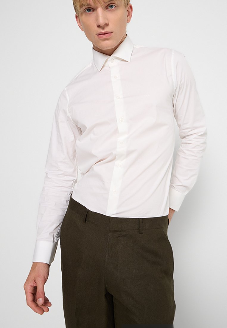 Blonde man in een wit overhemd met knopen en donkerbruin broek, staande met één hand in zijn zak tegen een effen achtergrond.