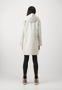 Lett, knelange regnjakke i lys grå med hette, elastisk liv, og lange ermer, kombinert med svarte leggings og joggesko.