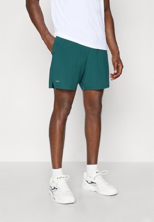 Shorts de sport pour homme en teal foncé avec fentes sur les côtés, fabriqués en tissu léger. Avec un logo contrastant près de l'ourlet. Associés à un t-shirt blanc.