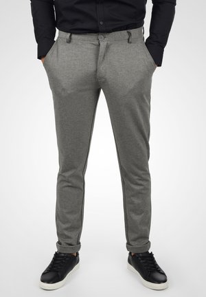 Homme portant un pantalon gris coupe slim retroussé aux chevilles, une chemise noire boutonnée, les mains dans les poches, et des baskets noires à semelles blanches.