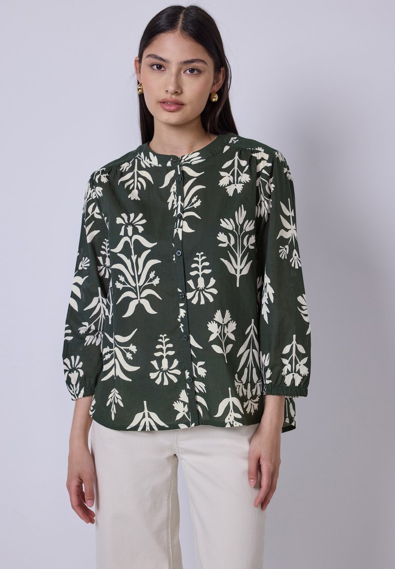 Blusa verde scuro con stampa botanica bianca. Il design include maniche a sbuffo a tre quarti e un collo rotondo con bottoni.