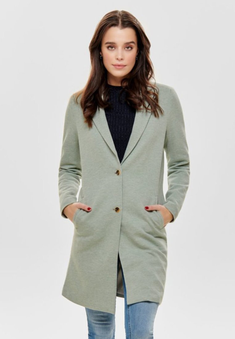 Blazer verde lungo con rever a scasso, due tasche frontali e bottoni marroni, realizzato in tessuto morbido, abbinato a un maglione scuro sotto.